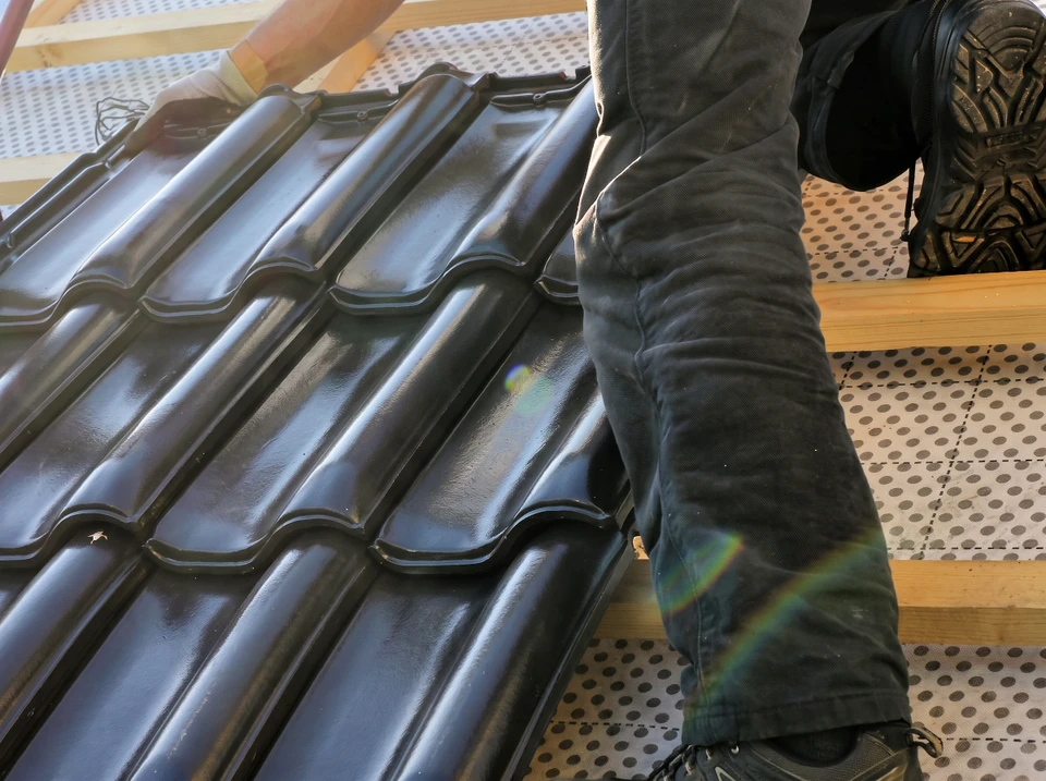 Couvreur en tenue de travail grise posant des tuiles métalliques noires brillantes sur une toiture en cours d'installation